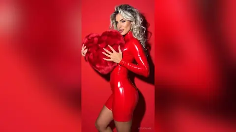 JessycaPage poses in a tight red latex dress while holding a large red furry heart against a red background.