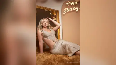 Glamorous blonde woman in a sparkling beige cutout dress posing in front of a Happy Birthday sign.