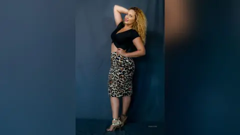 Smiling blonde woman in a black bodysuit and leopard print skirt posing against a blue studio backdrop.