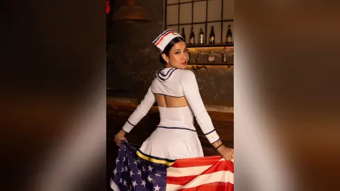 JuliahLewis poses in a white sailor-style outfit, looking back over her shoulder while holding an American flag behind her.