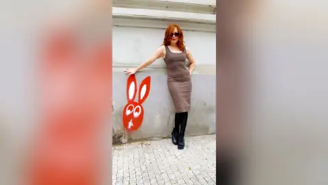 Redheaded woman in a brown midi dress and black boots posing by a red bunny street art mural.