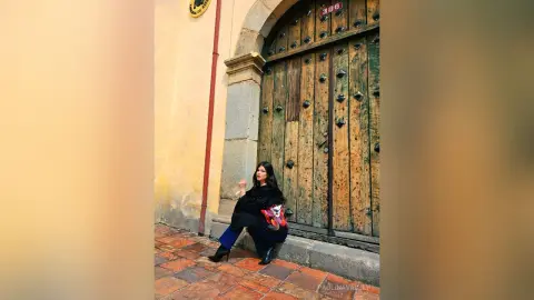 Woman in black top and blue jeans sitting by a rustic wooden door in a sunny cobblestone alleyway.