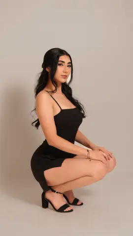 Young woman in a sleek black mini dress and high heels posing against a neutral studio background.
