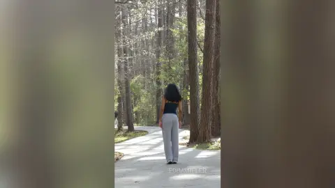 Woman in a black tank top and grey trousers walking down a peaceful forest path on a sunny day.