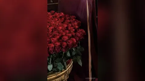 AlienaMoore — AlienaMoore stands next to a large gold vase overflowing with deep red roses.