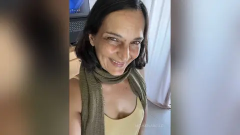 MichaelaBelle smiles at the camera wearing a tan tank top and an olive green scarf.