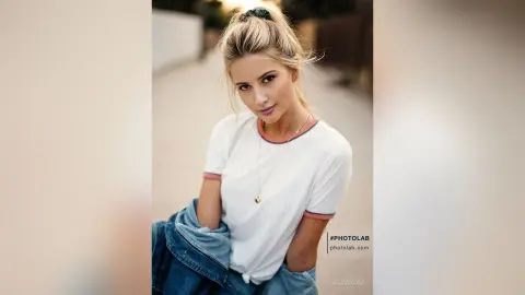 Blonde woman in a white t-shirt and denim jacket smiling in a soft outdoor setting.