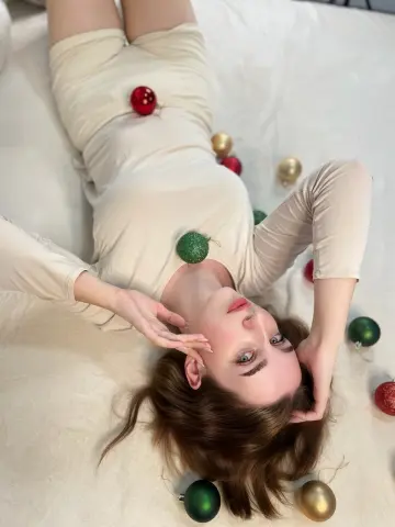 Young woman in a cream long-sleeve outfit lying on white bedding surrounded by colorful Christmas ornaments.