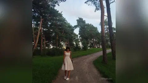 Young woman in a white floral sundress standing on a dirt path in a peaceful pine forest at dusk.