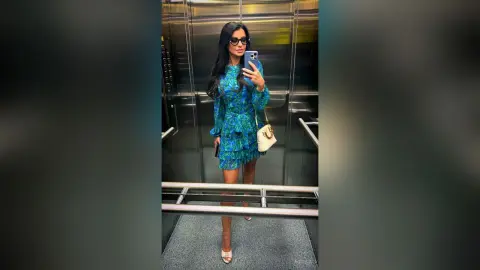 Stylish woman in a blue floral mini dress and sunglasses taking a mirror selfie in an elevator.