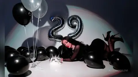 Glamorous woman in a black dress celebrating her 29th birthday with silver balloons and a cake in a moody setting.