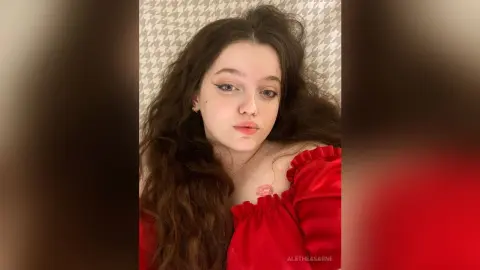 Young woman with long wavy hair wearing a red off-the-shoulder top in a soft, moody indoor setting.