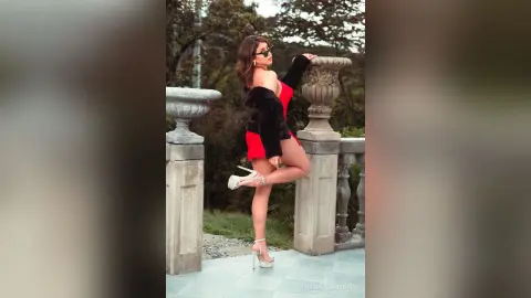 RebecaVillalobos stands on a stone balcony wearing a red bodysuit and black fur shrug with white high heels.