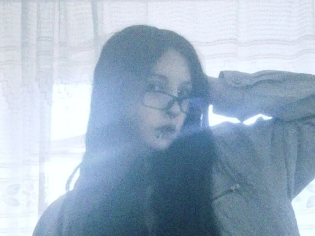Moody portrait of a woman with long dark hair and glasses wearing a casual jacket in a bright, hazy room.