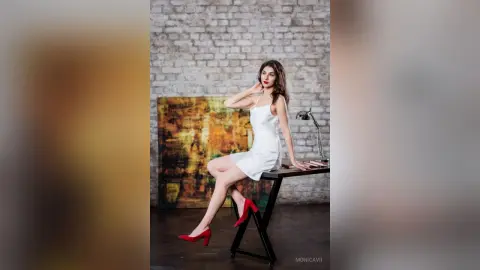 Elegant woman in a white cocktail dress and red heels sitting by an abstract painting in a studio setting.