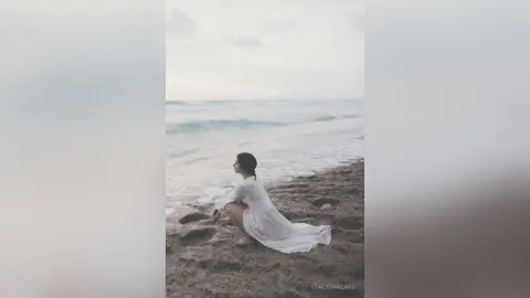 StacyHarland sits on a sandy beach wearing a long white dress against the ocean waves.