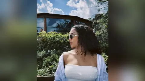 Young woman in a white tube top and sunglasses relaxing outdoors near lush green plants under a blue sky.