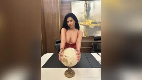 JasminGlance wears a low-cut red outfit while sitting at a desk and holding a decorative globe.