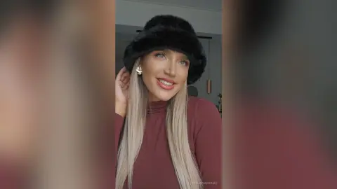 TiffanyAddams wearing a black furry hat and a maroon turtleneck, smiling with her long blonde hair down.