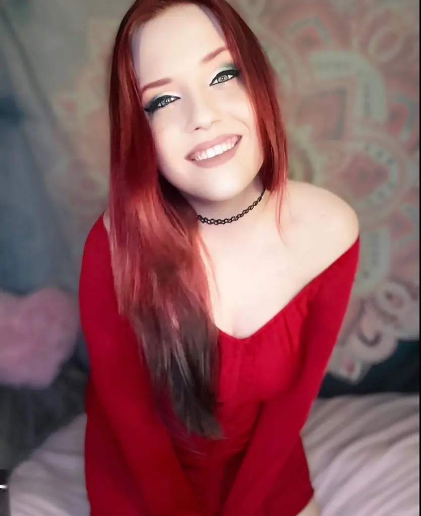 VioletOctober poses with long red hair, wearing a black top and a black choker with a silver bat pendant.