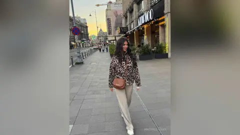 Smiling woman in leopard print jacket and beige trousers walking on a city street at sunset.