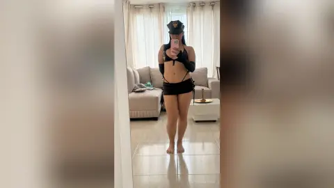 Woman in black police officer cosplay outfit taking a mirror selfie in a bright living room.