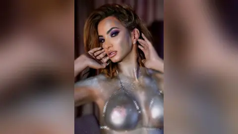 FelishaRose poses with her hands near her face, wearing silver glittery body paint and a thin silver chain around her neck.