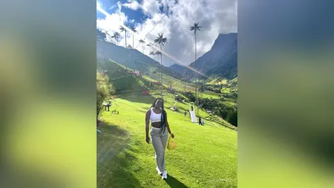 ValeryMei stands on a grassy hillside wearing a white tank top and light grey pants, surrounded by tall palm trees and mountains.