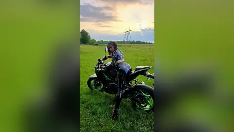 Stylish woman in black leather outfit sitting on a black motorcycle in a green field at sunset.