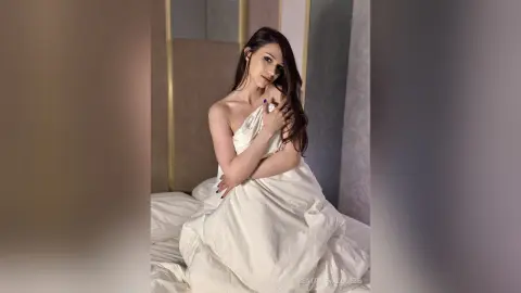 Elegant woman in white silk bedding posing gracefully in a bright, minimalist bedroom setting.