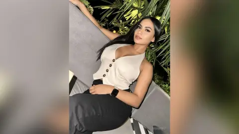 MiaKorz sits on a grey couch wearing a white sleeveless top and black pants, posing with her arm up against the backrest.