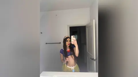 Woman in blue princess costume and gold crown taking a mirror selfie in a bright bathroom.