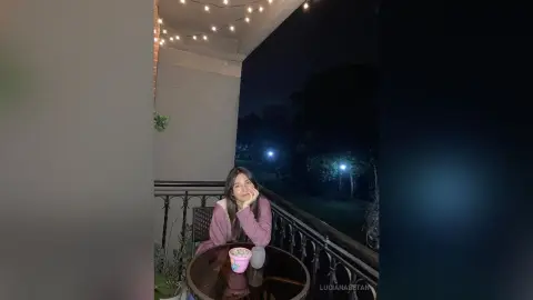 Relaxed woman in a mauve sweater sitting at a balcony table under fairy lights at night.