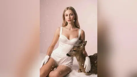 Blonde woman in a white satin bodysuit sitting in a dreamy snowy setting with a deer.
