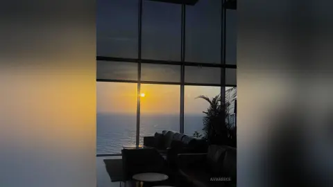 Golden sunset view over the ocean from a modern luxury penthouse living room with large floor-to-ceiling windows.