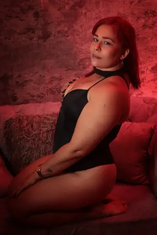 Red-haired woman in a black bodysuit posing on a velvet sofa in moody red lighting.
