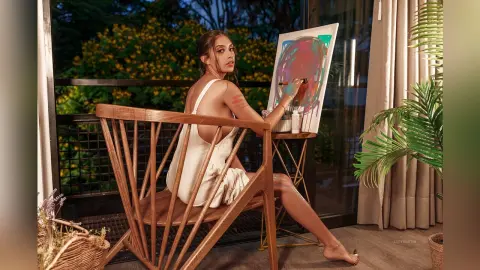 LucyMartin sits on a wooden chair in a white slip dress, holding a paintbrush next to a colorful canvas painting.