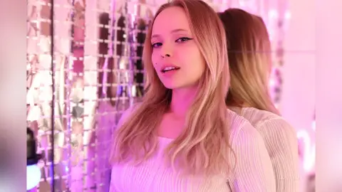 LucyLoy with long blonde hair and winged eyeliner poses in a white ribbed top against a shimmering pink background.