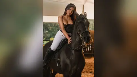 Elegant woman in white equestrian pants riding a black horse in a rustic outdoor stable setting.