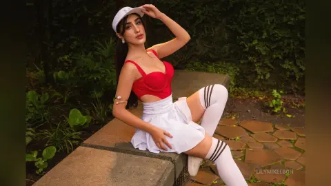 Young woman in a red crop top and white mini skirt posing outdoors in a lush green garden setting.
