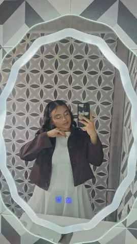 Young woman in a white dress and dark jacket taking a selfie in a modern bathroom with patterned tiles.