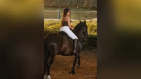 Elegant woman in white equestrian pants riding a black horse in a rustic outdoor paddock at sunset.