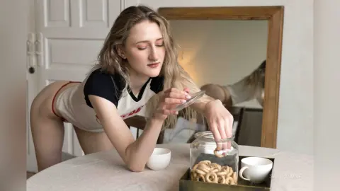 Blonde woman in a white t-shirt sitting at a dining table opening a glass jar of snacks in a bright room.