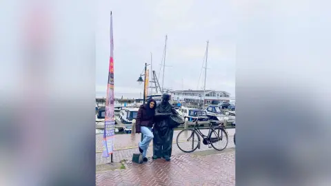 AnnaBanks stands outdoors near a harbor with boats and masts, wearing a dark jacket and blue jeans.
