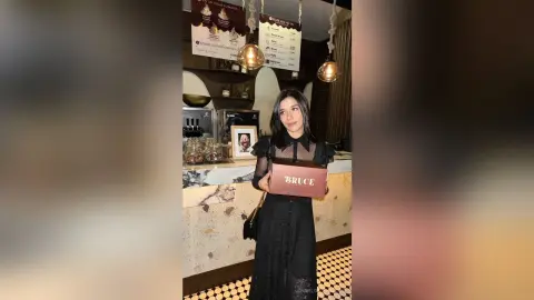 Young woman in a black dress holding a Bruce gift box in a cozy cafe setting.