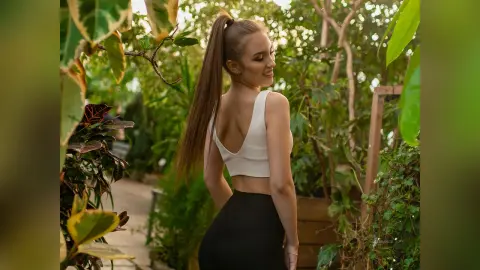 DelanaGrace poses outdoors in a white crop top, showing her back and long brown hair tied in a high ponytail.