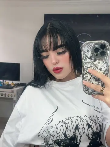 Young woman with black bangs in a white graphic t-shirt taking a casual mirror selfie indoors.