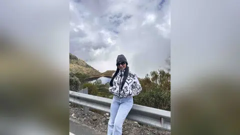 Woman in a black and white graphic sweatshirt and jeans standing by a scenic mountain lake under cloudy skies.