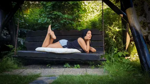 MissyJolie lies on her stomach on a wooden swing, wearing a black tank top and denim shorts with her feet up behind her.