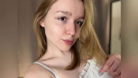 Blonde woman in a white lace camisole smiling in a bright, minimalist indoor setting.